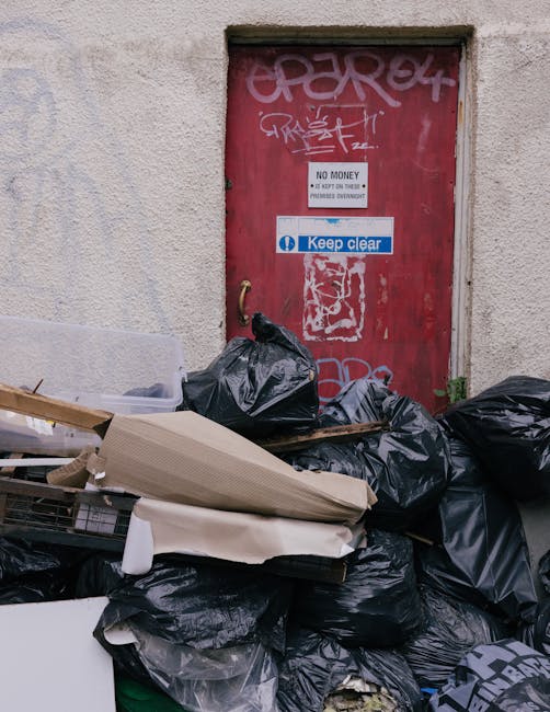 A middle-aged man with dark hair, wearing a black T-shirt with white text and dark trousers, is seen placing a white refuse sack into a large, cylindrical metal trash bin on a paved outdoor area. The 