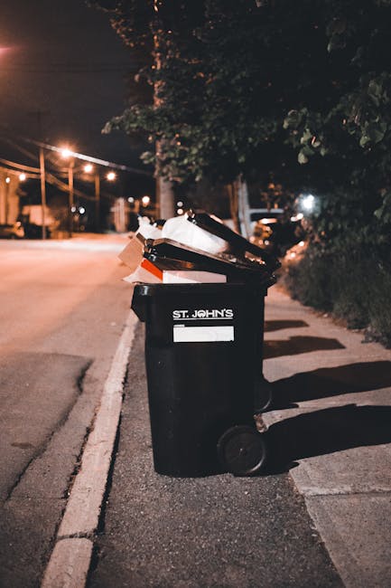 A large green wheelie bin filled with red and black bags of construction or landscaping materials, including flexible gravel or soil bags, positioned outside against a dark brick wall. The bin is moun