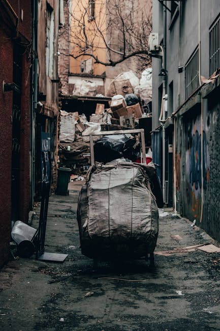 A narrow urban alleyway cluttered with large black and beige garbage bags, some torn open revealing waste inside, positioned on the uneven, stained asphalt ground. In the background, there are numerou