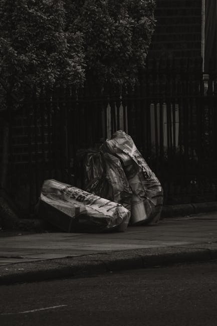 A narrow urban alleyway cluttered with large black and beige garbage bags, some torn open revealing waste inside, positioned on the uneven, stained asphalt ground. In the background, there are numerou